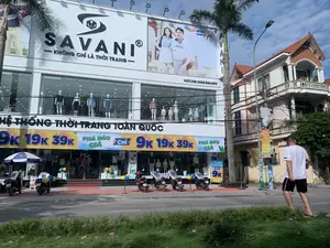Bối cảnh là mặt tiền của một cửa hàng thời trang lớn có tên là SAVANI, nằm trên một con phố. Tòa nhà cửa hàng có màu trắng, gồm nhiều tầng với mặt kính lớn. Trên cùng là biển hiệu lớn với logo và tên thương hiệu 'SAVANI', cùng khẩu hiệu 'KHÔNG CHỈ LÀ THỜI TRANG'. Bên cạnh là một tấm biển quảng cáo lớn có hình người mẫu nam nữ và dòng chữ 'EASY WEAR EASY LIFE', 'HOTLINE: 0888 566 599'. Phía trên tầng trệt có dòng chữ 'HỆ THỐNG THỜI TRANG TOÀN QUỐC'. Cửa hàng đang có chương trình khuyến mãi lớn, được thể hiện qua các băng rôn với nội dung 'PHÁ ĐẢO GIÁ', 'SALE HÈ RỰC CHÁY' và các mức giá hấp dẫn như 'TỪ 9k 19k 39k'. Bên trong cửa hàng, có thể thấy nhiều ma-nơ-canh đang trưng bày quần áo. Phía trước cửa hàng, một vài chiếc xe máy đang đỗ trên vỉa hè. Có một chiếc ô quảng cáo và các poster nhỏ. Một người đàn ông mặc áo phông trắng đang đi bộ ở lề đường. Cảnh quan xung quanh có cây cọ, cột đèn và một tòa nhà dân sinh bên cạnh. Một cột điện có treo biển hiệu với logo của ngân hàng AGRIBANK.。（日本語訳）。背景は、ある通りに面したSAVANIという名前の大きなファッション店の正面です。店舗の建物は白色で、何階建てかで、大きなガラス張りです。最上部には、「SAVANI」のロゴとブランド名、そして「KHÔNG CHỈ LÀ THỜI TRANG（ただのファッションではない）」というスローガンが書かれた大きな看板があります。その隣には、男女のモデルの写真と「EASY WEAR EASY LIFE」、「HOTLINE: 0888 566 599」という文字が書かれた大きな広告看板があります。1階の上には、「HỆ THỐNG THỜI TRANG TOÀN QUỐC（全国ファッションシステム）」という文字が書かれています。店は大規模なプロモーションキャンペーンを実施しており、それは「PHÁ ĐẢO GIÁ（価格破壊）」、「SALE HÈ RỰC CHÁY（燃えるような夏セール）」といった内容や、「9k、19k、39kから」といった魅力的な価格が書かれた横断幕で示されています。店内には、多くのマネキンが服を陳列しているのが見えます。店の前には、数台のバイクが歩道に停まっています。広告用のパラソルが1つと、小さなポスターがいくつかあります。白いTシャツを着た男性が一人、路肩を歩いています。周囲の景観には、ヤシの木、街灯、そして隣には住宅ビルがあります。一本の電柱には、AGRIBANK銀行のロゴが入った看板が掲げられています。