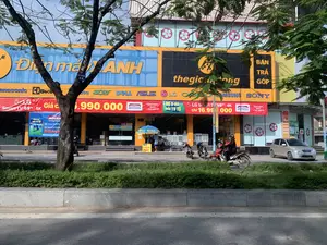 Bức ảnh chụp mặt tiền của một siêu thị điện máy lớn, có hai thương hiệu nổi bật là 'Điện máy XANH' và 'thegioididong'. Tòa nhà có màu sắc chủ đạo là xanh và vàng. Nhiều biển hiệu và băng rôn quảng cáo được treo lên, với các nội dung như: 'BÁN TRẢ GÓP', 'LG Smart TV 65" 4K chỉ 16.990.000', 'MÊ Ơ-RÔ, ĐỔI TV TO', 'TẶNG NGAY QUẠT + TRẢ GÓP 0%'. Các logo thương hiệu điện tử nổi tiếng như Panasonic, Electrolux, Acer, Dell, Asus, LG, Samsung, Sony cũng xuất hiện.。Về hoạt động, có một người đang lái xe máy màu đỏ trên đường. Phía trước cửa hàng, có hai chiếc xe máy khác đang đậu và một chiếc ô tô màu bạc. Một vài người đang đứng dưới một chiếc ô che màu xanh của một quầy dịch vụ nhỏ, trên đó có ghi 'ĐIỂM BÁN BẢO HIỂM XE MÁY' và 'TRẢ GÓP TV TỦ LẠNH MÁY GIẶT'. Cảnh vật bao gồm một con đường nhựa, một dải phân cách trồng cây xanh, và một cây phượng lớn có gắn biển 'CÂY PHƯỢNG 18'. Bên trong cửa hàng, có thể thấy các sản phẩm điện tử như TV và quạt điều hòa đang được trưng bày.。（日本語訳）。大きな家電量販店の正面を撮影した写真で、「Điện máy XANH」と「thegioididong」という2つの目立つブランドがあります。建物の主な色調は青と黄色です。多くの看板や広告バナーが掲げられており、「分割払い販売」、「LGスマートテレビ 65インチ 4K わずか16,990,000」、「ユーロに夢中、大きなテレビに買い替えよう」、「扇風機を即プレゼント＋分割払い金利0%」といった内容が書かれています。パナソニック、エレクトロラックス、エイサー、デル、エイスース、LG、サムスン、ソニーといった有名な電子機器ブランドのロゴも見られます。人の動きについては、道路上で赤いバイクを運転している人が一人います。店の前には、他に2台のバイクと1台の銀色の自動車が停まっています。小さなサービスカウンターの青いパラソルの下に数人が立っており、そこには「バイク保険販売所」および「テレビ・冷蔵庫・洗濯機 分割払い」と書かれています。風景には、アスファルトの道路、緑が植えられた中央分離帯、そして「ホウオウボク 18」という札が付けられた大きなホウオウボクの木が含まれています。店内では、テレビや冷風扇などの電子製品が展示されているのが見えます。