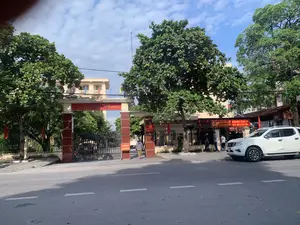 Bức ảnh chụp quang cảnh bên ngoài trụ sở Công an huyện Yên Định, tỉnh Thanh Hóa. Đây là một tòa nhà công vụ nằm bên một con đường nhựa.。Chữ viết trên các biển báo:。- Trên cổng chính có dòng chữ lớn màu vàng trên nền đỏ: 'CÔNG AN TỈNH THANH HÓA' và bên dưới là 'CÔNG AN HUYỆN YÊN ĐỊNH'.。- Bên phải cổng, có một biểu ngữ màu đỏ ghi: 'NƠI TIẾP NHẬN HỒ SƠ - ... - XỬ LÝ VI PHẠM GIAO THÔNG - TRẬT TỰ - ĐĂNG KÝ XE ÔTÔ, XE MÔTÔ'.。- Gần cổng có một biển báo di động màu đỏ với chữ trắng: 'YÊU CẦU XUỐNG XE XUẤT TRÌNH GIẤY TỜ'.。Hoạt động và cảnh vật:。- Địa điểm: Trụ sở công an cấp huyện.。- Hoạt động: Một người dân đang đi bộ vào cổng. Một nhóm người, bao gồm một cảnh sát mặc sắc phục màu xanh, đang đứng trên vỉa hè phía trước khu vực tiếp dân. Hoạt động chung có thể liên quan đến các thủ tục hành chính như đăng ký xe hoặc xử lý vi phạm giao thông.。- Vật thể: Một chiếc ô tô bán tải màu trắng đang đỗ ở lề đường. Bên trong sân có thể thấy một vài chiếc xe khác. Nhiều cây xanh lớn được trồng xung quanh khuôn viên. Bầu trời trong xanh, có mây trắng.。（日本語訳）。タインホア省イエンディン県警察本部の外観を撮影した写真。これはアスファルト道路の脇にある官公庁の建物です。看板の文字：。- 正門には、赤地に大きな黄色の文字で「CÔNG AN TỈNH THANH HÓA」（タインホア省警察）と書かれ、その下には「CÔNG AN HUYỆN YÊN ĐỊNH」（イエンディン県警察）と書かれています。- 門の右側には、赤い横断幕に「NƠI TIẾP NHẬN HỒ SƠ - ... - XỬ LÝ VI PHẠM GIAO THÔNG - TRẬT TỰ - ĐĂNG KÝ XE ÔTÔ, XE MÔTÔ」（書類受付場所 - ... - 交通違反・秩序違反処理 - 自動車・バイク登録）と書かれています。- 門の近くには、白文字の赤い移動式看板があり、「YÊU CẦU XUỐNG XE XUẤT TRÌNH GIẤY TỜ」（降車して書類を提示してください）と書かれています。活動と風景：。- 場所：県レベルの警察本部。- 活動：一人の住民が門に向かって歩いています。青い制服を着た警察官を含む一団が、住民受付エリアの前の歩道に立っています。一般的な活動は、車両登録や交通違反処理などの行政手続きに関連している可能性があります。- 物体：白いピックアップトラックが路肩に駐車しています。敷地内には他の車も数台見えます。敷地の周りには多くの大きな緑の木が植えられています。空は青く澄んでおり、白い雲があります。