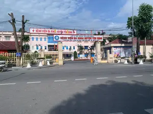 Bối cảnh là phía trước cổng chính của Bệnh viện Đa khoa huyện Yên Định, tỉnh Thanh Hóa. Tòa nhà bệnh viện nhiều tầng, sơn màu hồng, nằm ở phía sau cổng. Con đường phía trước được trải nhựa với vạch kẻ đường.。Các dòng chữ trên các biển hiệu có thể đọc được như sau:。- Biển hiệu lớn màu đỏ phía trên cổng: "+ BỆNH VIỆN ĐA KHOA HUYỆN YÊN ĐỊNH +"。- Biển hiệu trên trụ cổng: "SỞ Y TẾ TỈNH THANH HÓA", "BỆNH VIỆN ĐA KHOA HUYỆN YÊN ĐỊNH", địa chỉ "Xã Định Long - Huyện Yên Định - Tỉnh Thanh Hóa" và số điện thoại "0866 853-585".。- Tấm áp phích bên phải cổng thông báo: "KHÁM BỆNH BẢO HIỂM Y TẾ NGÀY CHỦ NHẬT" và liệt kê các chuyên khoa như Khoa Nội, Khoa Ngoại, Khoa Nhi, v.v.。Về hoạt động và các vật thể, một người mặc trang phục dài màu sáng và đội nón lá đang đi bộ ra khỏi cổng. Gần chốt bảo vệ bên phải, có hai người khác đang đứng, một trong số họ cũng đội nón lá. Một chiếc xe máy được đỗ ở lề đường bên trái. Khung cảnh được trang trí bằng các chậu cây cảnh lớn màu trắng, cây xanh và cờ phướn nhiều màu sắc treo phía trên.。（日本語訳）。背景は、タインホア省イエンディン県総合病院の正門前です。多層階建ての病院の建物はピンク色に塗られており、門の後ろに位置しています。正面の道路は舗装され、車線が引かれています。看板に書かれている文字は以下の通りです。- 門の上にある大きな赤い看板：「+ イエンディン県総合病院 +」。- 門柱の看板：「タインホア省保健局」、「イエンディン県総合病院」、住所「タインホア省イエンディン県ディンロン社」、電話番号「0866 853-585」。- 門の右側にあるポスターには、「日曜日の健康保険診療」と告知されており、内科、外科、小児科などの専門科がリストアップされています。人の動きや物体については、明るい色の長い服を着てノンラー（ベトナムの伝統的な笠）をかぶった人が門から歩いて出てきています。右側の警備員の詰所の近くに、他に2人が立っており、そのうちの1人もノンラーをかぶっています。左側の路肩にバイクが1台停められています。風景は、大きな白い観葉植物の鉢、緑の木々、そして上部に吊るされた色とりどりの幟旗で飾られています。