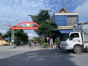 Bối cảnh là một con đường tại lối vào một khu dân cư ở Việt Nam, dưới bầu trời trong xanh có mây. Nổi bật trong ảnh là một chiếc cổng chào lớn màu đỏ bắc ngang đường, trên đó có dòng chữ vàng: "KHU DÂN CƯ TÂN NGƯ". Bên cạnh cổng, có một biển báo đường phố màu xanh ghi "ĐƯỜNG QUỐC LỘ 45" và một biển báo giao thông hình tròn viền đỏ, nền trắng có ký hiệu "6t", chỉ giới hạn trọng tải. Một chiếc xe tải màu trắng hiệu THACO FRONTIER đang ở trên đường, do một người đàn ông điều khiển. Hai người dân, trong đó có một phụ nữ đội nón lá, đang đạp xe đi vào khu dân cư. Bên lề đường có một chiếc xe máy màu đỏ đang đỗ và nhiều cây xanh.。（日本語訳）。背景は、雲のある青空の下、ベトナムのある住宅地の入り口にある道です。写真の中で目立っているのは、道をまたぐ大きな赤い歓迎ゲートで、その上には「KHU DÂN CƯ TÂN NGƯ」という黄色い文字があります。ゲートの隣には、「ĐƯỜNG QUỐC LỘ 45」と書かれた青い道路標識と、重量制限を示す「6t」という記号がある、赤い縁で白地の円形交通標識があります。THACO FRONTIERブランドの白いトラックが道路上におり、一人の男性が運転しています。二人の住民が、その中にはノンラーをかぶった女性もおり、自転車をこいで住宅地に入っていきます。道端には一台の赤いバイクが駐車しており、たくさんの緑の木々があります。