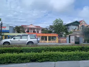 Bức ảnh chụp một con đường tại Thanh Hóa. Ở tiền cảnh là một chiếc ô tô SUV màu bạc và hàng cây bụi ven đường. Ở hậu cảnh là một tòa nhà hai tầng màu hồng, có vẻ là trụ sở của một công ty hoặc cơ quan nhà nước. Các văn bản có thể đọc được bao gồm:。Trên biển hiệu màu nâu ở cổng: 'CÔNG TY CỔ PHẦN XÂY DỰNG VÀ QUẢN LÝ GIAO THÔNG 1 ĐỊNH THANH HÓA' và 'FAX: 0373 51 24'.。Trên hai băng rôn màu đỏ treo trước tòa nhà: 'ĐẢNG CỘNG SẢN VIỆT NAM QUANG VINH MUÔN NĂM。' và 'TUÂN THỦ NỘI QUY, QUY TRÌNH AN TOÀN, VỆ SINH LAO ĐỘNG TẠI NƠI LÀM VIỆC.'.。Một phần của băng rôn màu vàng bị che khuất: 'CÔNG TY CỔ PHẦN XÂY DỰNG VÀ QUẢN LÝ GIAO TH... THAM GIA PHÒNG NGỪA, TỐ GIÁC CÁC HÀNH...'.。Bên trái, một biển hiệu màu xanh có chữ '...NGHIỆP'.。Khung cảnh cho thấy một con đường với các tòa nhà, bao gồm trụ sở công ty có cổng và bốt gác, cùng với các ngôi nhà dân. Hai lá cờ đỏ được treo trước tòa nhà chính. Không có hoạt động cụ thể nào của con người được ghi lại, ngoài sự hiện diện của một chiếc xe hơi trên đường.。（日本語訳）。写真はタインホア省のある道路を撮影したものです。後景にはピンク色の2階建ての建物があり、会社または国家機関の本社のようです。読み取れるテキストは以下の通りです。門にある茶色の看板には、「CÔNG TY CỔ PHẦN XÂY DỰNG VÀ QUẢN LÝ GIAO THÔNG 1 ĐỊNH THANH HÓA」（タインホア第1交通建設管理株式会社）と「FAX: 0373 51 24」。建物の前に掲げられた2つの赤い横断幕には、「ĐẢNG CỘNG SẢN VIỆT NAM QUANG VINH MUÔN NĂM。」（ベトナム共産党万歳。）と「TUÂN THỦ NỘI QUY, QUY TRÌNH AN TOÀN, VỆ SINH LAO ĐỘNG TẠI NƠI LÀM VIỆC.」（職場の内規、安全手順、労働衛生を遵守せよ）。一部が隠れている黄色の横断幕には、「CÔNG TY CỔ PHẦN XÂY DỰNG VÀ QUẢN LÝ GIAO TH... THAM GIA PHÒNG NGỪA, TỐ GIÁC CÁC HÀNH...」（交通建設管理株式会社... 行為の防止、告発に参加...）。左側には、「...NGHIỆP」という文字が書かれた青い看板があります。風景は、門と警備員詰所のある会社の本社を含む建物や民家が立ち並ぶ道路を示しています。2つの赤い旗が本館の前に掲げられています。道路上の車の存在以外に、特定の人の活動は記録されていません。