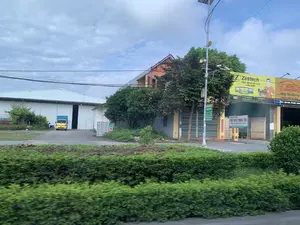 Đây là khung cảnh ven đường với nhiều tòa nhà và biển hiệu thương mại. Ở phía sau, có một nhà kho lớn màu trắng, trước cửa có một chiếc xe tải nhỏ màu vàng đang đỗ. Bên cạnh nhà kho là các tòa nhà dân sinh và cửa hàng. Nổi bật là các biển quảng cáo lớn. Một biển hiệu màu vàng của "Zestech - TRUNG TÂM NỘI THẤT XE HƠI HOÀNG DƯƠNG", quảng cáo "MÀN HÌNH Ô TÔ XUẤT MỸ". Một biển hiệu khác màu đỏ của "HOÀNG DƯƠNG" với khẩu hiệu "Nâng Tầm Dịch Vụ" và số hotline "0868 08 08 98". Một biển hiệu nhỏ hơn ghi địa chỉ có các từ "...ĐỊNH LONG - YÊN ĐỊNH - THANH...". Phía trước một ngôi nhà có cổng sắt, có một tấm biển trắng ghi "Chính Chủ CHO THUÊ PHÒNG TRỌ" với các tiện ích như "RỘNG RÃI, THOÁNG MÁT, CÓ WIFI, CÓ CHỖ ĐỂ Ô TÔ". Cảnh vật còn có cây xanh, cột điện gắn loa phát thanh công cộng, camera an ninh và tấm pin năng lượng mặt trời. Hàng cây bụi ở tiền cảnh bị mờ, cho thấy ảnh được chụp từ một phương tiện đang di chuyển.。（日本語訳）。これは多くの建物や商業看板がある道端の風景です。後方には大きな白い倉庫があり、その前には一台の黄色い小型トラックが停まっています。倉庫の隣には住宅や店舗があります。目立っているのは大きな広告看板です。一つは「Zestech - HOANG DUONG自動車内装センター」の黄色い看板で、「アメリカ輸出向けカーモニター」を宣伝しています。もう一つは「HOANG DUONG」の赤い看板で、「サービスのレベルを上げる」というスローガンとホットライン番号「0868 08 08 98」が書かれています。より小さな看板には「...DINH LONG - YEN DINH - THANH...」という言葉を含む住所が書かれています。鉄の門がある家の前には、「オーナー直貸し 貸室あり」と書かれた白い看板があり、「広々、風通し良好、WIFIあり、駐車スペースあり」といった設備が記載されています。風景には他に緑の木々、公共放送用のスピーカーが取り付けられた電柱、防犯カメラ、そして太陽光パネルがあります。前景の茂みの列はぼやけており、写真が移動中の乗り物から撮影されたことを示しています。