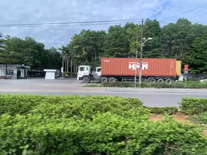 Hình ảnh chụp một chiếc xe đầu kéo màu trắng đang vận chuyển một thùng container lớn màu đỏ cam trên một con đường nhựa. Trên thành của thùng container có in chữ 'HMM' màu trắng và 'KOBC' màu đen. Bối cảnh cho thấy chiếc xe đang di chuyển ngang qua cổng vào của một khu vực có vẻ là khu công nghiệp hoặc nhà máy, có chốt bảo vệ và rào chắn. Xung quanh có nhiều cây xanh, cột điện, dây điện và camera an ninh được lắp trên cột. Ở tiền cảnh là một hàng rào cây xanh bị mờ do chuyển động, cho thấy bức ảnh được chụp từ một phương tiện đang di chuyển. Gần cổng có một biển báo giao thông hình tròn với số '5'.。（日本語訳）。白いトレーラーヘッドが、アスファルトの道路で大きなオレンジレッドのコンテナを運んでいる写真。コンテナの側面には、白い文字で「HMM」と黒い文字で「KOBC」が印刷されている。背景から、この車両が工業団地か工場と思われるエリアの入口ゲートを横切っていることがわかる。そこには警備員の詰め所と遮断機がある。周囲には多くの緑の木々、電柱、電線、そして柱に設置された防犯カメラがある。前景には、動きによってぼやけた生け垣があり、写真が移動中の乗り物から撮影されたことを示している。ゲートの近くには、数字の「5」が書かれた円形の交通標識がある。