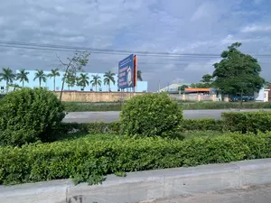 Bối cảnh là một con đường với cây xanh ở hai bên. Ở phía xa, có một tấm pano tuyên truyền lớn màu xanh và cam. Nội dung trên pano có thể đọc được là: "CHƯƠNG TRÌNH MỤC TIÊU QUỐC GIA - GIẢM NGHÈO BỀN VỮNG GIAI ĐOẠN 2021 - 2025". Phía sau tấm pano là một tòa nhà lớn màu xanh nhạt, có thể là một nhà máy hoặc nhà xưởng, với một hàng cây cọ được trồng phía trước. Có một số chữ viết trên tường của tòa nhà nhưng không thể đọc rõ. Ở phía xa bên phải, có một tòa nhà khác với một băng rôn màu đỏ treo phía trước, nhưng nội dung cũng không thể đọc được. Bầu trời nhiều mây. Cảnh vật cho thấy đây có thể là một khu công nghiệp hoặc khu vực ngoại ô.。（日本語訳）。背景は両側に緑の木々がある一本の道です。遠くには、青とオレンジ色の大きな宣伝看板があります。看板の内容は「国家目標プログラム - 持続可能な貧困削減 2021年～2025年」と読み取れます。看板の後ろには、水色の大きな建物があり、工場か作業場と思われ、その前にはヤシの木が植えられています。建物の壁にはいくつかの文字が書かれていますが、はっきりと読むことはできません。遠くの右側には、赤い横断幕が前に掲げられた別の建物がありますが、その内容も読み取れません。空は曇っています。この風景は、ここが工業団地か郊外の地域であることを示しています。