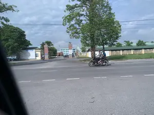 Hình ảnh chụp một con đường nhựa phía trước cổng vào của một công ty hoặc khu công nghiệp. Có một tấm biển thông báo lớn màu trắng ở bên trái cổng, trên đó có dòng chữ lớn có thể đọc được là "TUYỂN 300 LAO ĐỘNG". Các chi tiết khác trên biển báo không thể đọc rõ nhưng dường như là thông tin tuyển dụng. Trên cột cổng cũng có một biển báo nhỏ nhưng chữ viết không thể nhận dạng. Về hoạt động, hai người đàn ông đội mũ bảo hiểm đang đi trên một chiếc xe máy trên đường. Một người khác cũng đang đi xe máy gần cổng. Bên trong khuôn viên công ty có thể thấy một chiếc xe tải và các tòa nhà nhà xưởng. Khung cảnh có nhiều cây xanh và bầu trời nhiều mây.。（日本語訳）。ある会社または工業団地の入口ゲート前にあるアスファルトの道路を写した写真。ゲートの左側には大きな白い告知板があり、そこには「労働者300名募集」と読み取れる大きな文字が書かれている。看板の他の詳細ははっきりと読み取れないが、募集情報のようだ。ゲートの柱にも小さな看板があるが、文字は識別できない。人の動きとしては、ヘルメットをかぶった男性2人が道路でバイクに乗っている。別の人物もゲートの近くでバイクに乗っている。風景には緑の木々が多く、空は曇っている。