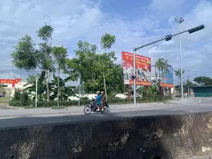 この写真はベトナムのある街の風景を撮影したものです。二人の女性がアスファルトの道路でバイクを運転しています。道端には緑の木々と大きなプロパガンダパネルがあります。赤いパネルが目立っており、そこには「国の刷新を祝う - 栄光ある党 - 2024年甲辰の新年を祝う」、「党を祝い、春を祝う」、そして「全人民が団結し、新しい農村と文明的な都市を建設する」という文言が書かれています。このパネルには、アグリバンク - イエンディン県支店のロゴもあります。左奥には、門と「...ディンリエン」および「文化・スポーツセンター」と書かれた赤い看板のある建物があります