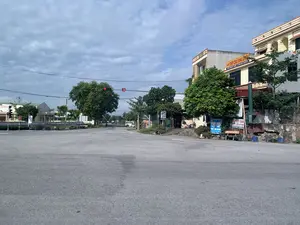 Bối cảnh là một ngã tư đường nhựa rộng rãi, khá vắng vẻ dưới bầu trời nhiều mây. Có một hệ thống đèn tín hiệu giao thông đang bật đèn đỏ và hiển thị đồng hồ đếm ngược. Bên phải đường là một tòa nhà nhiều tầng, hoạt động như một nhà hàng, với nhiều cây xanh xung quanh. Trên tòa nhà có các biển hiệu với nội dung:。1. Biển hiệu màu vàng: "QUÁN THỊT CHÓ LONG LUYẾN", bên dưới có thông tin "ĐC: NGÃ 4 NGA LIÊN - YÊN ĐỊNH - ĐT: 0345 730 818 - 0345 587 811". Trên mặt tiền tòa nhà có khắc số "2020".。2. Biển hiệu màu xanh dương: "Nhà Hàng Long Luyến", "THỊT CHÓ", "SỐNG - CHÍN ĐỦ MÓN", "Kính Chào Quý Khách。".。3. Một biển hiệu nhỏ khác cũng ghi "THỊT CHÓ", "SỐNG - CHÍN ĐỦ MÓN".。Phía xa bên trái có một tòa nhà công cộng với biển hiệu chỉ đọc được một phần là "...-THỂ THAO XÃ...". Trên con đường vắng, có một người đang lái xe máy ở phía xa.。（日本語訳）。背景は、曇り空の下、広々としてかなり閑散としたアスファルトの交差点です。赤信号が点灯し、カウントダウンタイマーが表示されている交通信号機があります。道路の右側には、レストランとして営業している多層階の建物があり、周りには多くの緑の木々があります。建物には以下の内容の看板があります：。1. 黄色い看板：「QUÁN THỊT CHÓ LONG LUYẾN」（ロン・ルエン犬肉店）、その下には「住所：ガーリエン交差点 - イエンディン - 電話：0345 730 818 - 0345 587 811」という情報があります。建物の正面には「2020」という数字が刻まれています。2. 青い看板：「Nhà Hàng Long Luyến」（ロン・ルエン レストラン）、「THỊT CHÓ」（犬肉）、「SỐNG - CHÍN ĐỦ MÓN」（生肉 - 調理済み各種料理）、「Kính Chào Quý Khách。」（お客様、ようこそ。）。3. 別の小さな看板にも「THỊT CHÓ」（犬肉）、「SỐNG - CHÍN ĐỦ MÓN」（生肉 - 調理済み各種料理）と書かれています。遠くの左側には公共の建物があり、看板には「...-THỂ THAO XÃ...」（...-村のスポーツ...）と一部しか読めない文字が書かれています。閑散とした道路の上では、遠くにバイクを運転している人が一人います。