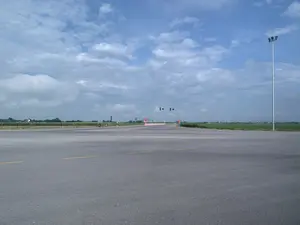 Bức ảnh chụp một ngã tư đường nhựa rộng lớn và vắng vẻ, có thể ở một khu vực ngoại ô hoặc nông thôn. Bầu trời xanh và có nhiều mây. Tại ngã tư có lắp đặt hệ thống đèn tín hiệu giao thông. Dọc bên đường có các biển báo giao thông, trong đó có thể nhận ra một biển báo "STOP" và một biển báo cấm đi ngược chiều. Con đường được trải nhựa với vạch kẻ đường màu vàng. Hai bên đường là những cánh đồng cây trồng xanh mướt. Nhìn ra xa là những ngôi nhà của khu dân cư. Phía bên phải có một cột đèn đường cao. Toàn bộ khung cảnh rất yên tĩnh và không có phương tiện giao thông nào đang di chuyển.。（日本語訳）。写真は、郊外または田舎にあると思われる、広くて閑散としたアスファルトの交差点を写しています。空は青く、雲が多いです。交差点には信号機システムが設置されています。道路沿いには交通標識があり、その中には「STOP」の標識と逆走禁止の標識が見えます。道路はアスファルトで舗装されており、黄色の車線が引かれています。道路の両側には、青々とした作物の畑が広がっています。遠くには住宅地の家々が見えます。右側には高い街灯が一本あります。全体的な風景は非常に静かで、走行中の車両は一台もありません。