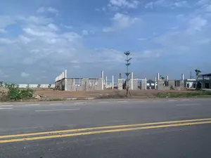 Bức ảnh chụp một công trường xây dựng nằm bên cạnh một con đường nhựa. Bầu trời xanh và có nhiều mây trắng. Con đường nhựa có vạch kẻ đôi liền nét màu vàng ở giữa. Tại công trường, nhiều ngôi nhà đang trong giai đoạn xây dựng dở dang, để lộ phần khung cột bê tông và các bức tường xây bằng gạch block. Một số giàn giáo bằng gỗ được dựng lên để hỗ trợ thi công. Mặt đất tại công trường là đất trống, màu nâu. Có một vài cây xanh nhỏ mới trồng và một số lùm cây, bao gồm cả cây chuối ở phía bên trái. Không có bất kỳ văn bản, biển báo nào có thể đọc được trong ảnh. Không có người hoặc hoạt động cụ thể nào được nhìn thấy, chỉ có cảnh quan của một dự án xây dựng đang tiến hành.。（日本語訳）。アスファルトの道路の隣にある建設現場を写した写真です。空は青く、白い雲がたくさんあります。アスファルトの道路の中央には、黄色の二重実線が引かれています。現場では、多くの家が建設途中の段階にあり、コンクリートの柱のフレームとブロックレンガで造られた壁がむき出しになっています。施工をサポートするために、いくつかの木製の足場が組まれています。現場の地面は茶色の更地です。新しく植えられた小さな緑の木が数本と、左側にあるバナナの木を含むいくつかの茂みがあります。写真には、読み取れる文字や看板は一切ありません。