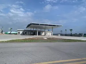 Hình ảnh cho thấy một công trình đang trong quá trình xây dựng, có khả năng là một trạm xăng hoặc trạm dừng nghỉ ven đường. Công trình bao gồm một tòa nhà một tầng bằng bê tông chưa được sơn và một mái che lớn được chống đỡ bởi các cột bê tông. Sân đã được đổ bê tông nhưng công trình có vẻ vẫn còn dang dở, với các vật liệu xây dựng như ván gỗ và một đống đồ được che bởi tấm bạt lớn màu xanh lá cây và trắng ở bên trái. Có một vài chiếc xe máy đậu dưới mái che. Xung quanh khu vực được bao bọc bởi một bức tường bê tông và có một số cây xanh mới được trồng. Phía trước là một con đường nhựa có vạch kẻ vàng. Bầu trời trong xanh và có mây. Không có biển hiệu hay chữ viết nào có thể đọc được trong ảnh.。（日本語訳）。画像は建設中の建物を示しており、ガソリンスタンドか道路沿いの休憩所である可能性があります。建物は、未塗装のコンクリート製の一階建ての建物と、コンクリートの柱で支えられた大きな屋根で構成されています。地面はコンクリートで舗装されていますが、工事はまだ未完成のようで、左側には木の板などの建設資材や、緑と白の大きな防水シートで覆われた物の山があります。屋根の下には数台のバイクが停まっています。周辺はコンクリートの壁で囲まれており、新しく植えられた木がいくつかあります。手前には黄色い線が引かれたアスファルトの道路があります。空は青く、雲があります。写真には読み取れる看板や文字はありません。