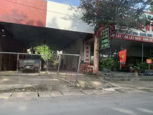 Bức ảnh chụp khung cảnh phía trước một cửa hàng kinh doanh vật liệu xây dựng ven đường. Cửa hàng có một khu vực nhà kho với mái che bằng khung thép, bên trong có một chiếc xe tải nhỏ đang đỗ. Một hàng rào lưới thép di động được đặt ở phía trước.。Bên phải là mặt tiền của cửa hàng, có treo một lá cờ Tổ quốc (cờ đỏ sao vàng). Nhiều chậu cây cảnh được đặt trên bậc thềm. Có nhiều biển hiệu quảng cáo với các nội dung có thể đọc được như sau:。- Biển hiệu dọc màu xanh lá cây: "ĐẠI HỒNG VẬT LIỆU XÂY DỰNG XI MĂNG SẮT THÉP".。- Biển hiệu ngang màu đỏ: "...VỆ SINH - GẠCH, NGÓI", "ỐP LÁT - ĐÁ LÁT NỀN - BỒN BỂ".。- Biển hiệu dọc màu trắng: "THIẾT BỊ VẬT LIỆU XÂY DỰNG".。- Biển hiệu nhỏ màu đỏ: "CHO THUÊ CỐP PHA ĐT: 0383 108 872 0904 787 459".。Ngoài ra, có thể thấy các chồng ngói lợp màu đỏ được xếp ở bên ngoài cửa hàng. Không có hoạt động mua bán nào rõ ràng đang diễn ra tại thời điểm chụp ảnh.。（日本語訳）。写真は、道端にある建材店の前の風景を撮影したものです。店には鉄骨の屋根がある倉庫エリアがあり、中には小型トラックが1台停まっています。正面には移動式の金網フェンスが設置されています。右側は店の正面で、国旗（金星紅旗）が掲げられています。多くの観葉植物の鉢が上がり口に置かれています。以下のように内容が読み取れる多くの広告看板があります。- 緑色の縦看板：「ダイホン建材 セメント 鉄鋼」。- 赤色の横看板：「...衛生 - レンガ、瓦」、「タイル張り - 床石 - タンク」。- 白色の縦看板：「建設資材設備」。- 赤色の小さな看板：「型枠レンタル 電話：0383 108 872 0904 787 459」。その他、店の外には赤い屋根瓦の山が積まれているのが見えます。撮影時点では、明確な売買活動は行われていないようです。