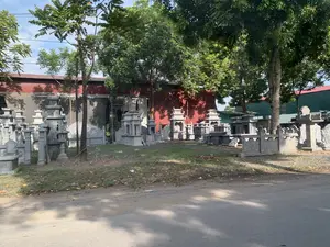 Đây là một khu vực ngoài trời, dường như là một xưởng chế tác hoặc nơi trưng bày các sản phẩm đá mỹ nghệ, có thể là một làng nghề đá ở Thanh Hóa. Có rất nhiều công trình bằng đá như lăng mộ đá, mộ đá, đèn đá, và lan can đá được xếp đặt trên một bãi đất cỏ. Các sản phẩm này có màu xám, được chạm khắc các hoa văn và chi tiết phức tạp. Trên một ngôi mộ đá ở phía bên trái, có một chữ Hán được khắc, trông giống chữ "福" (Phúc). Phía sau khu trưng bày là một nhà xưởng lớn với tường và mái tôn màu đỏ. Nhiều cây xanh mọc xen kẽ trong khu vực. Phía trước là một con đường trải nhựa. Khung cảnh tĩnh, không có hoạt động của con người được ghi lại rõ ràng, nhưng cho thấy đây là một cơ sở sản xuất và kinh doanh các sản phẩm từ đá.。（日本語訳）。ここは屋外のエリアで、美術石製品の工房または展示場のようであり、タインホア省の石工芸村かもしれません。石の陵墓、石の墓、石灯籠、石の欄干といった多くの石造りの建造物が、草地の上に並べられています。これらの製品は灰色で、複雑な模様やディテールが彫刻されています。左側にある石の墓の上には、漢字一文字が刻まれており、「福」（フック）の字のように見えます。展示エリアの後ろには、壁と屋根が赤いトタンでできた大きな工房があります。エリア内には多くの緑の木々が点在して生えています。前方にはアスファルトで舗装された道があります。光景は静かで、人間の活動ははっきりと記録されていませんが、ここが石製品を生産・販売する施設であることを示しています。