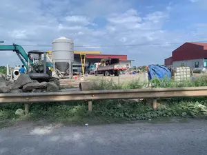 Bức ảnh chụp một khu vực trông giống như một công trường xây dựng hoặc một xưởng sản xuất vật liệu, nằm cạnh một con đường. Ở phía trước là một lan can đường bộ bằng kim loại. Bên trong khu vực công trường, có nhiều máy móc hạng nặng như một máy xúc màu xanh lá, một silo lớn màu xám dùng để chứa vật liệu, và một cấu trúc cần cẩu giàn màu vàng. Có một chiếc xe tải cẩu và các xe tải khác đang đỗ. Nhiều tảng đá lớn, ống cống bê tông, và các vật liệu xây dựng khác nằm rải rác trên mặt đất. Ở phía sau là các tòa nhà lớn kiểu nhà xưởng có mái tôn màu đỏ. Có một vài người đang làm việc, bao gồm một người đàn ông mặc áo xanh đứng gần máy xúc và những người khác đang thi công một công trình nhỏ hơn ở phía xa. Bầu trời trong xanh, có mây trắng. Có chữ viết trên cabin máy xúc nhưng không thể đọc rõ.。（日本語訳）。手前には金属製のガードレールがあります。現場内には、緑色のショベルカー、資材を貯蔵するための大きな灰色のサイロ、そして黄色のガントリークレーン構造物のような多くの重機があります。クレーン付きトラックやその他のトラックが駐車しています。大きな岩、コンクリートの土管、その他の建設資材が地面に散らばっています。背景には、赤いトタン屋根の大きな工場風の建物があります。数人が作業しており、その中にはショベルカーの近くに立っている青いシャツの男性や、遠くでより小さな構造物を建設している他の人々が含まれています。空は青く澄んでおり、白い雲があります。ショベルカーの運転台には文字が書かれていますが、はっきりと読むことはできません。