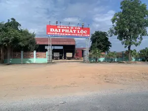 Bức ảnh chụp lối vào một gara sửa chữa ô tô có tên "Đại Phát Lộc". Phía trên cổng vào có một tấm biển hiệu lớn màu đỏ với các thông tin chi tiết. Nội dung trên biển hiệu được ghi lại như sau: "GARA Ô TÔ ĐẠI PHÁT LỘC. CHUYÊN SỬA CHỮA MÁY, GẦM, MÁY CÔNG TRÌNH - CƠ KHÍ: TIỆN, PHAY, BÀO - BÁN PHỤ TÙNG. ĐC: DỐC LÊ - YÊN THÁI - YÊN ĐỊNH - THANH HÓA. ĐT: 0917 981 078 - 0965 781 078".。Loại hình địa điểm là một gara ô tô, chuyên về xe tải và máy công trình. Bên trong sân, có thể thấy hai chiếc xe tải ben đang đỗ trong một nhà xưởng có mái tôn. Khu vực được bao quanh bởi hàng rào và có trồng một số cây xanh. Con đường phía trước gara là đường đất đá. Bầu trời có mây.。（日本語訳）。「ダイ・ファット・ロック」という名前の自動車修理ガレージの入り口を撮影した写真です。入り口の上には、詳細情報が書かれた大きな赤い看板があります。看板の内容は次の通りです：「ダイ・ファット・ロック自動車ガレージ。専門：エンジン、足回り、建設機械の修理 - 機械加工：旋盤、フライス、平削り - 部品販売。住所：タインホア省イエンディン県イエンタイ社ゾックレー。電話：0917 981 078 - 0965 781 078」。この場所の種類は自動車ガレージで、トラックと建設機械を専門としています。敷地内には、トタン屋根の作業場に2台のダンプトラックが駐車しているのが見えます。このエリアはフェンスで囲まれており、いくつかの木が植えられています。ガレージの前の道は未舗装の砂利道です。空は曇っています。