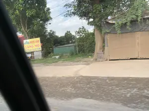 Bức ảnh chụp cảnh ven đường, có thể được chụp từ bên trong một chiếc ô tô. Nổi bật là một tấm biển quảng cáo màu vàng của một cơ sở sản xuất có tên "NGỌC LÂM". Nội dung trên biển ghi rõ: "Cơ sở NGỌC LÂM, Chuyên LĂNG MỘ, TÂM TƯỜNG RÀO - HOA VĂN XÂY DỰNG". Điều này cho thấy đây là một xưởng chuyên chế tác lăng mộ, tường rào và các chi tiết hoa văn bằng đá hoặc xi măng cho công trình xây dựng. Bên cạnh tấm biển có các tấm vật liệu xây dựng (có thể là các bộ phận của lăng mộ hoặc hàng rào) được xếp chồng lên nhau. Phía sau là cây cối, một nhà kho lợp tôn màu xanh và một công trình tạm có tường bằng ván và mái che bạt. Không có hoạt động nào của con người được ghi lại.。（日本語訳）。写真は道端の風景を撮影したもので、車内から撮影された可能性があります。目立っているのは、「NGỌC LÂM」という名前の製造施設の黄色い看板です。看板には「NGỌC LÂM施設、専門：陵墓、フェンス・塀 - 建築用装飾模様」と明記されています。これは、この施設が建設工事用に、石やセメントで陵墓、塀、装飾模様のディテールを専門に製作する工房であることを示しています。看板の隣には、建設資材の板（陵墓やフェンスの部品である可能性があります）が積み重ねられています。後方には木々、青いトタン屋根の倉庫、そして板壁と防水シートの屋根を持つ仮設の建物があります。人間の活動は記録されていません。