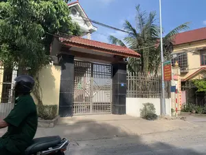 Bối cảnh là một con đường trong khu dân cư ở Việt Nam vào một ngày nắng đẹp. Ở phía trước bên trái, có một người đang lái xe máy, mặc trang phục màu xanh lá cây và đội mũ bảo hiểm. Phía sau là những ngôi nhà dân, trong đó nổi bật là một ngôi nhà có cổng lớn với hai trụ ốp đá hoa cương màu đen và mái che lợp ngói đỏ. Cánh cổng làm bằng sắt. Trên trụ cổng có gắn biển số nhà màu xanh ghi số "01". Ở bên phải, trên một cột điện, có treo một tấm biển tuyên truyền với nội dung: "TÔI VÀ BẠN HÃY CHUNG TAY VÌ MỘT MÔI TRƯỜNG XANH, SẠCH, ĐẸP CỦA CHÚNG TA。". Xung quanh có nhiều cây xanh, bao gồm cả cây dừa, và nhiều dây điện, dây cáp vắt ngang trên bầu trời trong xanh.。（日本語訳）。背景は、晴れた日のベトナムの住宅街の道です。左手前には、緑色の服を着てヘルメットをかぶった人がバイクを運転しています。後ろには民家が並んでおり、その中でも目立つのは、黒い花崗岩で覆われた2本の柱と赤い瓦で葺かれた屋根を持つ大きな門のある家です。門は鉄製です。門柱には「01」と書かれた青い番地プレートが取り付けられています。右側には、電柱に「私とあなたは、私たちの緑豊かで清潔で美しい環境のために手を取り合いましょう。」という内容のプロパガンダ看板が掲げられています。周りにはココナッツの木を含む多くの緑の木々があり、青い空には多くの電線やケーブルが横切っています。