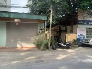 Bối cảnh là một con đường tại một khu dân cư. Bên trái là một ngôi nhà có cửa cuốn màu xám đang đóng kín, phía trên mái hiên bằng tôn màu xanh có treo một chiếc đèn lồng kiểu cũ. Ở phía bên phải, có một cửa hàng với các biển hiệu quảng cáo sơn. Các dòng chữ có thể đọc được bao gồm: 'ĐẠI LÝ CHỈNH SÂM', 'SƠN CAO CẤP CÔNG NGHỆ NANO', 'NANO TREND', và số điện thoại '0978 066 182'. Một biển số nhà màu xanh ghi số '511' được gắn trên tường. Đỗ trên vỉa hè là một chiếc xe máy màu đen, và phía sau có thể thấy một phần đầu của một chiếc ô tô. Cảnh vật còn có cây xanh, một vài chậu cây cảnh, và một cột điện với hệ thống dây điện chằng chịt.。（日本語訳）。背景は、ある住宅街の道です。左側には灰色のシャッターが閉まっている家があり、青いトタンの庇の上には古いスタイルの提灯が吊るされています。右側には、ペンキの広告看板がある店があります。読み取れる文字には、「ĐẠI LÝ CHỈNH SÂM」、「SƠN CAO CẤP CÔNG NGHỆ NANO」、「NANO TREND」、そして電話番号「0978 066 182」が含まれます。壁には「511」と書かれた青い番地プレートが取り付けられています。歩道には黒いバイクが停まっており、その後ろには自動車の先端部分が見えます。風景には他に、緑の木々、いくつかの鉢植え、そして複雑に絡み合った電線のある電柱があります。