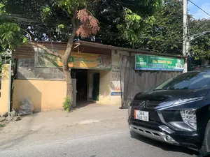 Bối cảnh là một con đường ở Thanh Hóa. Hình ảnh cho thấy một ngôi nhà cấp bốn, tường sơn màu vàng, có vẻ là một cửa hàng hoặc đại lý phân phối. Phía trước ngôi nhà có nhiều cây xanh và hệ thống dây điện chằng chịt. Một chiếc ô tô màu đen đang ở bên phải đường, che một phần mặt tiền cửa hàng.。Các thông tin văn bản có thể đọc được trên các biển hiệu:。1. Biển quảng cáo màu vàng của Fami: "THÊM LỰA CHỌN GIÁ KHÔNG ĐỔI* *SO VỚI FAMI NGUYÊN VỊ".。2. Biển hiệu màu xanh lá cây của công ty bia: "CÔNG TY CỔ PHẦN BIA HÀ NỘI - THANH HÒA", "Bia THANH HOA", "HABECO", "THABREW SILVER". Địa chỉ: "Đ/C: 152 Quang Trung - Phường Ngọc Trạo - TP. Thanh Hóa", Điện thoại: "02373.857.541 - 02373.752.699", Website: "www.biathanhhoa.com.vn".。3. Biển hiệu cũ đã mờ: "Linh", "Bán buôn, b...", có thể là "TẠP HÓA". Địa chỉ: "ĐC: Phù Hưng 3, Yên Thái - Yên...", Điện thoại: "01202 105 861".。Không có hoạt động rõ ràng của con người trong ảnh, nhưng các biển hiệu cho thấy đây là một địa điểm kinh doanh, có thể là bán buôn và bán lẻ các mặt hàng tiêu dùng.。（日本語訳）。背景はタインホア省のある道路です。画像には、壁が黄色に塗られた平屋の家が写っており、店舗または代理店のようです。家の前には多くの緑の木々と、複雑に絡み合った電線があります。黒い車が道路の右側にあり、店の正面の一部を覆っています。看板から読み取れるテキスト情報：。1. Famiの黄色い広告看板：「選択肢が増えても価格はそのまま* *FAMIオリジナルテイストと比較」。2. ビール会社の緑色の看板：「ハノイ-タインホアビール株式会社」、「タインホアビール」、「HABECO」、「THABREW SILVER」。住所：「タインホア市ゴックチャオ坊クアンチュン通り152番地」、電話番号：「02373.857.541 - 02373.752.699」、ウェブサイト：「www.biathanhhoa.com.vn」。3. 色褪せた古い看板：「Linh」、「卸売、b...」、おそらく「雑貨店」。住所：「フー・フン3、イエン・タイ - イエン...」、電話番号：「01202 105 861」。写真には明確な人の活動は見られませんが、看板からここが、おそらく消費財の卸売および小売を行う商業施設であることがわかります。