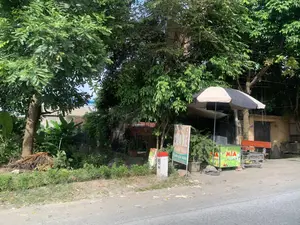 Các dòng chữ ghi trên các biển hiệu và vật thể trong ảnh:。- Biển hiệu màu trắng: "RỬA XE", "THAY DẦU", "Nước giải khát", "CÓ ĐÁ VIÊN".。- Xe đẩy màu xanh: "Nước MÍA siêu sạch".。- Cột mốc đường: "H5", "44".。Đây là hình ảnh một quán nhỏ ven đường, có thể ở khu vực nông thôn hoặc ngoại ô. Quán được che bởi một chiếc ô lớn màu đen và trắng. Theo các biển hiệu, nơi này cung cấp dịch vụ rửa xe, thay dầu và bán nước giải khát, cụ thể là nước mía "siêu sạch" được đặt trên một chiếc xe đẩy màu xanh. Có một chiếc ghế gỗ dài để khách ngồi. Cảnh vật xung quanh có nhiều cây cối xanh tốt, phía sau có thể thấy nhà dân lợp mái ngói và một vài cây chuối. Bên lề đường có một cột mốc sơn trắng, đỉnh đỏ, ghi "H5" và "44". Không có hoạt động mua bán nào đang diễn ra và không có người trong ảnh.。（日本語訳）。写真の中の看板や物体に書かれている文字：。- 白い看板：「洗車」、「オイル交換」、「清涼飲料水」、「氷あり」。- 青いカート：「超清潔サトウキビジュース」。- 道路のキロポスト：「H5」、「44」。これは、田舎または郊外にある可能性のある、道端の小さな店の写真です。店は白と黒の大きな傘で覆われています。看板によると、この場所は洗車、オイル交換、清涼飲料水の販売サービスを提供しており、具体的には青いカートの上に置かれた「超清潔」なサトウキビジュースがあります。客が座るための長い木製の椅子があります。周囲の風景には緑豊かな木々が多く、後ろには瓦屋根の民家と数本のバナナの木が見えます。道端には、白く塗られ、上部が赤く、「H5」と「44」と書かれたキロポストがあります。売買の活動は行われておらず、写真には誰もいません。