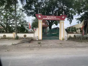 Bức ảnh chụp cổng vào của một khu tưởng niệm liệt sĩ. Trên vòm cổng màu đỏ có dòng chữ trắng: "NHÀ BIA TƯỞNG NIỆM CÁC ANH HÙNG LIỆT SỸ XÃ YÊN PHONG". Cổng sắt màu xanh lá cây đang đóng. Bên trong khuôn viên, có thể thấy một công trình kiến trúc với mái ngói, trên đó có tấm biển ghi "TỔ QUỐC GHI CÔNG". Phía bên trái cổng có một cột cờ treo lá cờ Tổ quốc Việt Nam (cờ đỏ sao vàng). Khu vực được bao quanh bởi tường rào và có nhiều cây xanh. Không có người hay hoạt động nào diễn ra trong ảnh.。（日本語訳）。ある烈士記念地区の入り口ゲートを撮影した写真。赤いアーチゲートの上には白い文字で「NHÀ BIA TƯỞNG NIỆM CÁC ANH HÙNG LIỆT SỸ XÃ YÊN PHONG」（イエンフォン社英雄烈士記念碑）と書かれている。緑色の鉄の門は閉まっている。敷地内には、瓦屋根の建築物が見え、その上には「TỔ QUỐC GHI CÔNG」（祖国は功績を記す）と書かれたプレートがある。門の左側には、ベトナム国旗（金星紅旗）を掲げた旗竿がある。この区域は塀で囲まれており、多くの緑の木々がある。写真の中には人や活動は見られない。