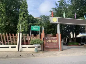 Đây là hình ảnh chụp bên ngoài cổng vào của Công sở xã Yên Phong. Trên cột cổng xây bằng gạch có một tấm biển màu nâu ghi chữ 'CÔNG SỞ XÃ YÊN PHONG' và số điện thoại 'ĐT 037 3 843 320'. Bên cạnh đó, có một biển báo màu xanh của 'NGÂN HÀNG CHÍNH SÁCH XÃ HỘI HUYỆN YÊN ĐỊNH', thông báo đây là 'ĐIỂM GIAO DỊCH XÃ YÊN PHONG' với 'Thời gian giao dịch: Từ 8 giờ 00 đến 12 giờ 00, ngày 6 hàng tháng'. Khuôn viên được bao quanh bởi hàng rào sắt màu nâu và có nhiều cây xanh. Phía trên cổng có cắm một số lá cờ nhỏ nhiều màu. Bên trong sân có một vài chiếc xe máy và một chiếc ô tô đang đỗ. Khung cảnh cho thấy đây là một trụ sở hành chính cấp xã.。（日本語訳）。これはイエンフォン村役場の入口ゲートの外側から撮影された写真です。レンガ造りの門柱には、「CÔNG SỞ XÃ YÊN PHONG」という文字と「ĐT 037 3 843 320」という電話番号が書かれた茶色い看板があります。その隣には、「NGÂN HÀNG CHÍNH SÁCH XÃ HỘI HUYỆN YÊN ĐỊNH」の青い看板があり、ここが「ĐIỂM GIAO DỊCH XÃ YÊN PHONG」であること、そして「取引時間：毎月6日、8時00分から12時00分まで」であることが告知されています。敷地は茶色い鉄柵で囲まれており、緑の木々がたくさんあります。門の上には、色とりどりの小さな旗がいくつか掲げられています。敷地内には、数台のバイクと1台の自動車が駐車しています。この光景は、ここが村レベルの行政庁舎であることを示しています。