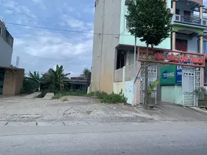 Bức ảnh chụp một con đường ở khu vực dân cư tại Việt Nam. Bên phải là một tòa nhà nhiều tầng, nơi có một quán karaoke. Phía trên cổng vào có một biển hiệu lớn màu đỏ với dòng chữ 'KÍNH CHÀO QUÝ KHÁCH' và một biển hiệu khác ghi 'KARAOKE 666'. Bên dưới là một biển quảng cáo lớn của hãng bia 'TUBORG' với dòng chữ 'VỊ BIA TUYỆT HẢO TỪ 1880', cùng với tên quán có thể là 'KARAOKE THA...' và số điện thoại 'ĐT: 0976 8...' nhưng không đầy đủ. Cổng sắt vào quán đang mở. Trên một cây cột gần đó có gắn biển số nhà '29'. Bên trái tòa nhà là một khu đất trống, nền xi măng đã xuống cấp, phía sau có một ngôi nhà nhỏ hơn và nhiều cây cối, bao gồm cả cây chuối. Ở phía xa có một cột tháp viễn thông. Bầu trời có mây. Không có hoạt động nào của con người được ghi lại trong ảnh.。（日本語訳）。写真はベトナムの住宅街にある道を写したものです。右側には多階建ての建物があり、そこにはカラオケ店があります。入口の門の上には、「KÍNH CHÀO QUÝ KHÁCH」という文字が書かれた大きな赤い看板と、「KARAOKE 666」と書かれた別の看板があります。その下には、「VỊ BIA TUYỆT HẢO TỪ 1880」というキャッチコピーが入ったビールブランド「TUBORG」の大きな広告看板があり、店の名前は「KARAOKE THA...」で、電話番号は「ĐT: 0976 8...」とありますが、完全ではありません。店への鉄の門は開いています。近くの柱には「29」という番地のプレートが取り付けられています。建物の左側には空き地があり、セメントの地面は劣化しています。その後ろにはより小さな家と、バナナの木を含む多くの木々があります。遠くには通信塔があります。空は曇っています。写真には人間の活動は記録されていません。