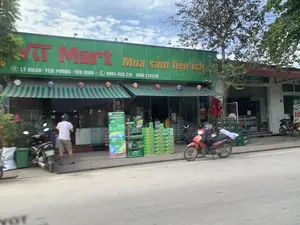 Bức ảnh chụp mặt tiền của một cửa hàng tiện lợi có tên 'VT Mart' nằm bên đường. Trên tấm biển hiệu lớn màu xanh lá cây có ghi dòng chữ 'VT Mart', 'Mua sắm tiện ích', địa chỉ 'LÝ NHÂN - YÊN PHONG - YÊN ĐỊNH' và số điện thoại '0964 550 219 - 0989 270 638'. Phía dưới biển hiệu là mái hiên sọc xanh trắng. Trước cửa hàng có một người đàn ông đang đứng, và một vài chiếc xe máy đang đỗ. Nhiều thùng hàng hóa, có vẻ là đồ uống, được xếp chồng lên nhau để trưng bày bên ngoài. Một lá cờ Việt Nam được treo ở phía trong. Khung cảnh cho thấy một con đường nhựa, cây cối và bầu trời có mây.。（日本語訳）。道路脇にある「VT Mart」という名前のコンビニエンスストアの正面を撮影した写真。大きな緑色の看板には、「VT Mart」、「便利な買い物」という文字、住所「LÝ NHÂN - YÊN PHONG - YÊN ĐỊNH」、そして電話番号「0964 550 219 - 0989 270 638」が書かれています。看板の下には、緑と白の縞模様の庇があります。店の前には男性が一人立っており、数台のバイクが停まっています。飲み物と思われる多くの商品箱が、屋外に陳列するために積み重ねられています。店内にはベトナムの国旗が掲げられています。