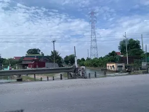 Bức ảnh chụp một khung cảnh nông thôn với một con đường nhựa và lan can bảo vệ ở phía trước. Bên cạnh đường là một con kênh hoặc mương thủy lợi, nước đang chảy xiết. Có một công trình nhỏ màu vàng, có thể là trạm bơm hoặc cống điều tiết nước, nằm bên bờ kênh. Xung quanh là nhà dân với mái ngói đỏ, cây cối um tùm, bao gồm cả các bụi chuối. Một người đang ngồi bên bờ kênh, gần đó có một chiếc xe đạp. Ở phía xa, một cột điện cao thế lớn nổi bật trên nền trời nhiều mây. Có một cột mốc bê tông bên phải ghi chữ "G12". Có một tấm biển quảng cáo và dòng chữ trên tòa nhà nhỏ nhưng không thể đọc rõ nội dung.。（日本語訳）。写真は、手前にアスファルトの道路とガードレールがある田舎の風景を写しています。道路の隣には水路または灌漑用の溝があり、水が勢いよく流れています。水路の岸には、ポンプ場か水門と思われる黄色い小さな建造物があります。周りには赤い瓦屋根の民家や、バナナの茂みを含む木々が鬱蒼と茂っています。一人の人が水路の岸に座っており、その近くに自転車が一台あります。遠くには、曇り空を背景に大きな高圧電線の鉄塔が目立っています。右側には「G12」と書かれたコンクリートの標柱があります。小さな建物には広告看板と文字がありますが、内容ははっきりと読み取れません。