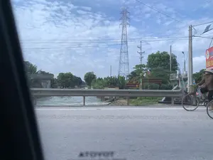 Bức ảnh ghi lại một khung cảnh ven đường ở một khu vực có vẻ là nông thôn hoặc ngoại ô. Ở phía trước là một con đường nhựa, được ngăn cách với một con kênh bằng một hàng rào chắn bằng kim loại. Dòng nước trên kênh đang chảy. Ở bên phải, một người phụ nữ đội nón lá đang đi xe đạp trên đường, trong giỏ xe có đựng rau. Phía sau hàng rào chắn là cây cối, bao gồm cả cây chuối, và một vài ngôi nhà. Nổi bật trên nền trời là một cột điện cao thế lớn và các cột điện nhỏ hơn. Có một tấm biển quảng cáo màu đỏ ở phía bên phải, có thể đọc được một phần chữ: 'HIỆU GÀ...', 'CHUYÊN...'. Bầu trời trong xanh và có nhiều mây.。（日本語訳）。写真は、田舎か郊外と思われる地域の道端の風景を捉えています。手前にはアスファルトの道があり、金属製のガードレールで水路と隔てられています。水路の水は流れています。右側では、ノンラーをかぶった女性が道を自転車で走っており、かごには野菜が入っています。ガードレールの後ろには、バナナの木を含む木々と、数軒の家があります。空を背景に、大きな高圧電線の鉄塔と、より小さな電柱が際立っています。右側には赤い広告看板があり、「HIỆU GÀ...」、「CHUYÊN...」という文字の一部が読み取れます。空は青く澄んでおり、雲が多くあります。