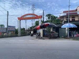 Hình ảnh ghi lại một con đường ở lối vào một ngôi làng tại Việt Nam, có một chiếc cổng chào lớn màu đỏ. Trên cổng có dòng chữ 'ĐƯỜNG KÊNH TÂY, THÔN LÝ NHÂN'. Phía dưới là biểu ngữ '“PHÁT HUY TRUYỀN THỐNG QUÊ HƯƠNG - THÔN LÝ NHÂN QUYẾT TÂM HOÀN THÀNH MỤC TIÊU XÂY DỰNG THÔN KIỂU MẪU”'. Hai bên trụ cổng có các khẩu hiệu 'KINH TẾ GIA ĐÌNH PHÁT TRIỂN' và 'XÃ HỘI...'.。Bên lề đường, một quầy hàng rong được dựng lên, sử dụng một chiếc xe ba gác (xe lôi) để bán các loại trái cây như chôm chôm hoặc vải, được che dưới những chiếc ô lớn. Một trong những chiếc ô có in thương hiệu 'Thạch Bích'. Một người đàn ông đang lái xe máy đi qua cổng làng. Phía sau có thể thấy nhà dân, cây cối và một cột điện cao thế. Dọc đường có nhiều biển quảng cáo cho các dịch vụ địa phương như 'NHÀ HÀNG HIỀN THANH' chuyên 'DÊ NÚI - CÁ SÔNG - GÀ ĐỒI', 'TRUNG TÂM CHĂM SÓC XE HƠI RỬA XE', và 'Đại Lý CHÈ THÁI NGUYỄN'.。（日本語訳）。画像はベトナムのある村の入り口の道を捉えたもので、大きな赤い歓迎ゲートがあります。ゲートには「ĐƯỜNG KÊNH TÂY, THÔN LÝ NHÂN」という文字が書かれています。その下には「故郷の伝統を発揮し、リーニャン村は模範的な村を建設するという目標を達成することを決意する」という横断幕が掲げられています。ゲートの柱の両側には「家族経済の発展」と「社会…」というスローガンがあります。道端には、三輪車（セーロイ）を使い、大きな傘の下でランブータンやライチなどの果物を売る露店が設けられています。傘の一つには「Thạch Bích」というブランド名が印刷されています。一人の男性がバイクを運転して村のゲートを通り過ぎています。後方には民家、木々、そして高圧電線の鉄塔が見えます。道沿いには、「山羊肉 - 川魚 - 地鶏」を専門とする「NHÀ HÀNG HIỀN THANH」、「TRUNG TÂM CHĂM SÓC XE HƠI RỬA XE」（自動車ケアセンター 洗車）、そして「Đại Lý CHÈ THÁI NGUYỄN」（タイグエン茶代理店）など、地元のサービスに関する多くの広告看板があります。