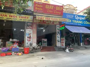 Bức ảnh chụp một dãy các cửa hàng liền kề nhau trên một con phố. Bên trái là cửa hàng bán đồ nhựa và nội thất với biển hiệu màu vàng ghi 'NHỰA NỘI THẤT', 'BÀN HỌC - KỆ GIÁ SÁCH'. Phía trước cửa hàng bày bán nhiều đồ nhựa gia dụng như chậu, rổ, và một chiếc xô màu đỏ. Ở giữa là 'CỬA HÀNG Tuấn Xuân' chuyên bán 'GIÀY - DÉP Thời Trang', 'GIÀY - DÉP DA', 'VÍ DA DÂY LƯNG'. Biển hiệu màu đỏ có ghi địa chỉ 'Đ/c: Phố Kiểu - Yên Trường - Yên Định - Thanh Hóa'. Một chiếc xe đạp màu trắng đang dựng trước cửa. Bên phải là 'SHOP Quỳnh' bán 'THỜI TRANG CÔNG SỞ' với biển hiệu màu xanh, địa chỉ tương tự. Bên trong cửa hàng có các ma-nơ-canh đang trưng bày quần áo. Một chiếc xe máy điện màu đỏ đang đỗ trên vỉa hè gần đó.。（日本語訳）。写真には、ある通りに隣り合って並ぶ一連の店舗が写っている。左側はプラスチック製品と家具を販売する店で、黄色い看板には「NHỰA NỘI THẤT」、「BÀN HỌC - KỆ GIÁ SÁCH」と書かれている。店の前には、たらい、かご、そして赤いバケツのような多くの家庭用プラスチック製品が陳列販売されている。中央は「CỬA HÀNG Tuấn Xuân」で、「GIÀY - DÉP Thời Trang」、「GIÀY - DÉP DA」、「VÍ DA DÂY LƯNG」を専門に販売している。赤い看板には「Đ/c: Phố Kiểu - Yên Trường - Yên Định - Thanh Hóa」という住所が書かれている。店の前に白い自転車が一台立てかけられている。右側は「SHOP Quỳnh」で、「THỜI TRANG CÔNG SỞ」を販売しており、青い看板で、住所は同様である。店内にはマネキンが衣類を陳列している。近くの歩道に赤い電動バイクが一台停まっている。