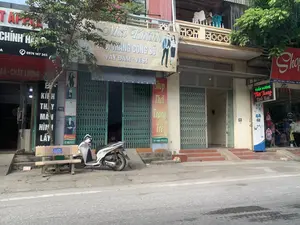 Bối cảnh là một con phố ở Việt Nam với nhiều cửa hàng san sát nhau, có thể ở một thị trấn thuộc huyện Yên Định, tỉnh Thanh Hóa. Bên lề đường có một chiếc xe máy màu trắng đang đỗ cạnh một chiếc ghế dài. Hầu hết các cửa hàng đều đóng cửa bằng cửa xếp sắt, nhưng một cửa hàng thời trang ở phía bên phải đang mở cửa và có người bên trong. Trên ban công tầng hai của một ngôi nhà, có quần áo đang được phơi.。Thông tin văn bản trên các biển hiệu có thể đọc được bao gồm:。1. Cửa hàng bên trái (một phần bị cây che khuất): Biển hiệu có chữ "...T APPLE", "CHÍNH HÃNG" và số điện thoại "0974 147 345". Biển dọc quảng cáo dịch vụ "ÉP KÍNH THAY MÀN HÌNH LẤY...". Số nhà là 67.。2. Cửa hàng ở giữa: Biển hiệu lớn ghi "Mai Khánh", chuyên "THỜI TRANG CÔNG SỞ", "VÁY ĐẦM - VEST". Địa chỉ và số điện thoại trên biển là "...Phố Kiều - Yên Trường - Yên Định - Thanh Hóa" và "ĐT: 0985 976 613". Bên cạnh có biển dọc "Shop Thời Trang Trẻ".。3. Cửa hàng bên phải: Một cửa hàng đang mở cửa với biển hiệu đèn LED "TUẤN HƯƠNG Thời Trang Trẻ Em". Một biển hiệu khác ở trên ghi địa chỉ "Đ/C: Phố Kiều - Yên Trường - Yên Định". Các số nhà 69 và 71 cũng được nhìn thấy.。（日本語訳）。背景はベトナムのある通りで、多くの店が隣接しており、おそらくタインホア省イエンディン県のある町です。道端には、長椅子の隣に一台の白いバイクが停まっています。ほとんどの店は鉄製のシャッターで閉まっていますが、右側にある一軒のファッション店は開いており、中に人がいます。ある家の2階のバルコニーには、服が干されています。読み取れる看板の文字情報には、以下のものが含まれます。1. 左側の店（一部が木で隠れている）：看板には「...T APPLE」、「CHÍNH HÃNG」（正規品）の文字と電話番号「0974 147 345」があります。縦長の看板には「ÉP KÍNH THAY MÀN HÌNH LẤY...」（ガラス圧着、画面交換、即時...）というサービスが広告されています。番地は67です。2. 中央の店：大きな看板には「Mai Khánh」と書かれており、「THỜI TRANG CÔNG SỞ」（オフィスファッション）、「VÁY ĐẦM - VEST」（ドレス - ベスト）を専門としています。看板にある住所と電話番号は「...Phố Kiều - Yên Trường - Yên Định - Thanh Hóa」（...キエウ通り - イエンチュオン - イエンディン - タインホア）と「ĐT: 0985 976 613」です。隣には「Shop Thời Trang Trẻ」（ヤングファッションショップ）という縦長の看板があります。3. 右側の店：開いている店で、「TUẤN HƯƠNG Thời Trang Trẻ Em」（トゥアンフオン 子供服）というLED看板があります。上にある別の看板には住所「Đ/C: Phố Kiều - Yên Trường - Yên Định」（住所：キエウ通り - イエンチュオン - イエンディン）が書かれています。69番と71番の番地も見えます。