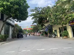 Bối cảnh là một con đường phố ở Việt Nam, có thể tại Thanh Hóa. Hai bên đường là những ngôi nhà cao tầng san sát nhau, tầng trệt được sử dụng làm cửa hàng kinh doanh. Cây xanh được trồng dọc hai bên đường, và có nhiều dây điện ở trên cao.。**Thông tin chữ viết:**。- Băng rôn đỏ treo ngang đường: "NHIỆT LIỆT CHÀO MỪNG KỶ NIỆM 70 NĂM CHIẾN THẮNG ĐIỆN BIÊN PHỦ (7/5/1954 - 7/5/2024)".。- Biển hiệu cửa hàng bên phải (tòa nhà màu vàng): "Huy Huấn ĐỒNG HỒ Đ/C: PHỐ KIỂU - YÊN...". Đây là một cửa hàng đồng hồ.。- Biển hiệu cửa hàng bên trái (tòa nhà màu trắng): "THÚY VÂN" (bị che khuất một phần).。- Các biển hiệu khác ở phía xa: "Bang ĐỒ CHƠI TRẺ EM PHOTO COPY IN VI TÍNH EP PLASTIC", "Tóc", "CH DINH".。**Cảnh vật và hoạt động:**。- **Loại địa điểm:** Một con phố với các cửa hàng và nhà dân.。- **Hoạt động cụ thể:** Người dân đang di chuyển trên đường bằng xe máy và xe đạp. Các cửa hàng hai bên đường đang mở cửa hoạt động.。- **Vật thể trong ảnh:** Nhiều xe máy đang di chuyển và đậu trên vỉa hè, một chiếc ô tô màu đen đang đỗ, các tòa nhà cao tầng, cây xanh, và một người phụ nữ đội nón lá đang đi xe ở phía xa.。（日本語訳）。背景はベトナムのある通りで、おそらくタインホア省でしょう。通りの両側には高層住宅が密集しており、1階は店舗として使用されています。街路樹が通りの両側に植えられており、上空には多くの電線があります。**書かれている文字の情報：**。- 通りを横切って掲げられた赤い横断幕：「ディエンビエンフーの戦い70周年記念（1954年5月7日 - 2024年5月7日）を熱烈に歓迎します」。- 右側の店の看板（黄色い建物）：「Huy Huấn 時計 住所：KIỂU通り - YÊN...」。これは時計店です。- 左側の店の看板（白い建物）：「THÚY VÂN」（一部が隠れている）。- 遠くにある他の看板：「Bang 子供のおもちゃ フォトコピー パソコン印刷 プラスチックラミネート」、「髪」、「CH DINH」。**風景と活動：**。- **場所の種類：**店舗や民家が並ぶ通り。- **具体的な活動：**人々がバイクや自転車で道路を移動しています。両側の店は営業しています。- **写真の中の物体：**多くのバイクが移動中であったり、歩道に駐車されていたりします。1台の黒い自動車が駐車しています。高層ビル、街路樹、そして遠くでノンラーをかぶった女性が乗り物に乗っています。