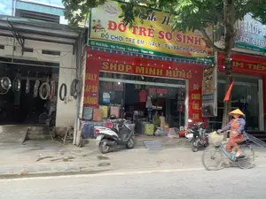 Đây là khung cảnh một con phố với các cửa hàng kinh doanh liền kề. Nổi bật ở trung tâm là 'Shop Minh Hưng', một cửa hàng có mặt tiền màu đỏ. Biển hiệu lớn màu vàng phía trên ghi rõ các thông tin:。- Tên cửa hàng và mặt hàng chính: 'Minh Hưng ĐỒ TRẺ SƠ SINH'。- Các sản phẩm khác: 'ĐỒ CHƠI TRẺ EM - VALY - TÚI XÁCH CÁC LOẠI'。- Địa chỉ: 'Phố Kiểu - Yên Trường - Yên Định - Thanh Hóa'。- Số điện thoại: '0392 963 886'。Cửa hàng này chuyên bán đồ dùng cho trẻ sơ sinh, đồ chơi, vali, túi xách. Phía trước cửa hàng có trưng bày nhiều vali và một vài chiếc xe máy đang đỗ.。Bên trái Shop Minh Hưng là một cửa hàng có vẻ như là tiệm sửa chữa hoặc bán phụ tùng xe, với nhiều vành xe và lốp xe được treo bên ngoài.。Bên phải là một cửa hàng khác có biển hiệu ghi '...LI-BALO-...' và tên cửa hàng '...IM TIẾN'.。Trên lòng đường, một người phụ nữ đội nón lá đang đi xe đạp. Cảnh vật cho thấy một ngày sinh hoạt bình thường trên một con phố thương mại.。（日本語訳）。これは、商店が隣接して営業している通りの風景です。中央で目立っているのは「Shop Minh Hưng」という、正面が赤い店です。上部にある大きな黄色い看板には、以下の情報が明記されています。- 店名と主要商品：「Minh Hưng 新生児用品」。- その他の商品：「子供のおもちゃ - スーツケース - 各種ハンドバッグ」。- 住所：「タインホア省イエンディン県イエンチュオン社キエウ通り」。- 電話番号：「0392 963 886」。この店は新生児用品、おもちゃ、スーツケース、ハンドバッグを専門に販売しています。店の前には多くのスーツケースが陳列されており、数台のバイクが停まっています。Shop Minh Hưngの左側には、修理店か自動車部品店のようで、多くのホイールリムとタイヤが外に吊るされています。右側には、看板に「...LI-BALO-...」と書かれ、店名が「...IM TIẾN」である別の店があります。路上では、ノンラーをかぶった女性が自転車に乗っています。この光景は、商店街でのありふれた日常の一日を示しています。