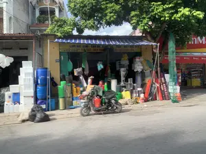 Trên biển hiệu màu vàng của cửa hàng có ghi các mặt hàng kinh doanh: "LƯỚI BẮT QUÁI - CAN ĐỰNG HŨ - VỎ - CHAI - MÁY SỤC OXY", "BỂ - NI LÔNG...V.V..", "BẠT - DÂY THỪNG" và số điện thoại. Biển số nhà là "121". Cửa hàng bên cạnh có biển hiệu "WinMart".。Đây là một cửa hàng mặt phố chuyên bán các loại vật tư, dụng cụ phục vụ cho việc đánh bắt và nuôi trồng thủy sản. Hàng hóa được trưng bày ra cả vỉa hè, bao gồm nhiều thùng xốp trắng, thùng phuy nhựa màu xanh, can nhựa, lồng bát quái, dây thừng và các vật dụng khác. Một chiếc xe máy điện đang đỗ ở phía trước. Bên cạnh là một cửa hàng tiện lợi WinMart. Khung cảnh cho thấy một con phố buôn bán vào ban ngày, dưới bóng một cây lớn.。（日本語訳）。店の黄色い看板には、取り扱い品目：「八卦網 - 壺・容器・瓶を入れる缶 - 酸素供給機」、「水槽 - ナイロン...など」、「シート - ロープ」と電話番号が書かれている。番地は「121」である。隣の店には「WinMart」の看板がある。これは、漁業や水産養殖用の各種資材や道具を専門に販売する路面店である。商品は歩道にまで陳列されており、多くの白い発泡スチロール箱、青いプラスチックドラム缶、プラスチック缶、八卦網、ロープ、その他の道具が含まれている。前には電動バイクが1台停まっている。隣にはコンビニエンスストアのWinMartがある。この光景は、大きな木の陰にある、日中の商店街の様子を示している。