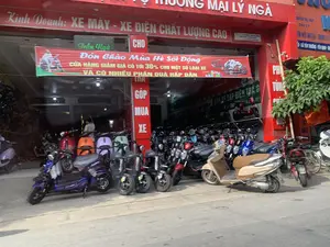 Đây là hình ảnh chụp mặt tiền của một cửa hàng kinh doanh xe máy và xe điện có tên "THƯƠNG MẠI LÝ NGÀ". Cửa hàng có mặt tiền lớn màu đỏ, trưng bày rất nhiều xe máy và xe điện mới, đủ màu sắc, ở cả bên trong và trên vỉa hè. Một băng rôn lớn màu đỏ được treo để quảng cáo chương trình khuyến mãi "Đón Chào Mùa Hè Sôi Động", với nội dung "CỬA HÀNG GIẢM GIÁ CÓ TỚI 30% CHO MỘT SỐ LOẠI XE VÀ CÓ NHIỀU PHẦN QUÀ HẤP DẪN". Các biển hiệu khác cũng cho thấy cửa hàng kinh doanh "XE MÁY - XE ĐIỆN CHẤT LƯỢNG CAO" và có hỗ trợ "TRẢ GÓP MUA XE". Một biển hiệu bên cạnh ghi địa chỉ tại "XÃ YÊN TRƯỜNG - YÊN ĐỊNH - THANH HÓA". Có một người đang ở bên trong cửa hàng. Phía bên phải có cây xanh và một lá cờ Việt Nam.。（日本語訳）。これは「THƯƠNG MẠI LÝ NGÀ」という名前のバイク・電動バイク販売店の正面を撮影した写真です。店は大きな赤い正面を持ち、店内と歩道に色とりどりの新しいバイクや電動バイクを多数展示しています。「活気ある夏を迎えよう」というプロモーションプログラムを宣伝するために大きな赤い横断幕が掲げられており、その内容は「一部の車種を最大30%割引、魅力的なプレゼント多数」です。他の看板も、店が「高品質バイク・電動バイク」を販売し、「分割払い購入」をサポートしていることを示しています。隣の看板には「タインホア省イエンディン県イエンチュオン社」という住所が書かれています。店内には一人の人がいます。右側には緑の木とベトナムの国旗があります。