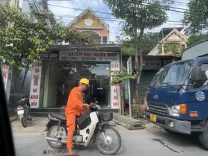 Bức ảnh chụp lại một cảnh đường phố ở Việt Nam. Ở vị trí trung tâm, một người đàn ông mặc bộ đồ bảo hộ lao động màu cam và đội mũ bảo hộ màu vàng đang ngồi trên một chiếc xe máy. Bên phải anh là một chiếc xe tải màu xanh dương, trên thân xe có ghi '6.5 TON'. Hai bên đường là các cửa hàng san sát. Bên trái là một cửa hàng có mặt tiền bằng kính với biển hiệu lớn ghi 'DƯƠNG ĐỊNH' và 'Chuyên QUẦN ÁO - GIẦY DÉP, DÂY LƯNG - VÍ DA SIN - ĐỒ SƠ SINH', cùng với số điện thoại 'ĐT: 0766 120 789 - 0766 124 789'. Một biển hiệu dọc khác ghi 'QUẦN ÁO TRẺ EM SƠ SINH BỈM' và số nhà '153'. Phía sau chiếc xe tải, có thể thấy một cửa hàng khác với biển hiệu 'Điện Tiệp ẢNH CHÂN DUNG INH LẤY NHANH' và số điện thoại. Khung cảnh cho thấy đường phố có cây xanh, nhiều dây điện chằng chịt phía trên, và các ngôi nhà dân. Một ngôi nhà ở phía sau có ghi năm '2012' trên phần mái.。（日本語訳）。ベトナムのある通りの風景を写した写真です。中央には、オレンジ色の作業着を着て黄色いヘルメットをかぶった男性がバイクに座っています。彼の右側には青いトラックがあり、車体には「6.5 TON」と書かれています。道路の両側には店が立ち並んでいます。左側にはガラス張りの店があり、「DƯƠNG ĐỊNH」と「専門 衣類 - 靴、ベルト - 革財布 - 新生児用品」と書かれた大きな看板と、電話番号「ĐT: 0766 120 789 - 0766 124 789」が記されています。別の縦長の看板には「子供服 新生児 おむつ」と書かれ、番地の「153」が記されています。トラックの後ろには、「Điện Tiệp ポートレート写真 スピードプリント」と電話番号が書かれた看板のある別の店が見えます。後ろにある家の一軒は、屋根の部分に「2012」年と書かれています。