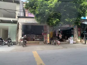 Đây là khung cảnh một con phố với các cửa hàng liền kề. Ở trung tâm là một cửa hàng có biển hiệu đã cũ ghi "THAO SACH MỚI CŨ ĐỒ DÙNG", có thể là một hiệu sách và bán đồ dùng văn phòng phẩm. Số nhà trên tường là "175". Bên cạnh, tại số nhà "177", là một cửa hàng khác, có vẻ là cửa hàng tạp hóa, với nhiều hàng hóa được xếp trong các thùng và bao tải phía trước. Một biển hiệu khác có chữ "SÁCH NGUYỄN". Một cây xanh lớn có tán lá sum suê che khuất một phần mặt tiền của các cửa hàng. Một vài chiếc xe máy và một chiếc xe đạp đang đậu trên vỉa hè. Con đường nhựa có vạch kẻ liền màu vàng. Bên trong cửa hàng số 175 có thể thấy các chồng sách và một tủ kính trưng bày.。（日本語訳）。これは、店舗が隣接する通りの光景です。中央には「THAO SACH MỚI CŨ ĐỒ DÙNG」と書かれた古い看板のある店があり、書店兼文房具店かもしれません。壁にある番地は「175」です。隣の番地「177」には別の店があり、雑貨店のようで、店の前には多くの商品が箱や袋に入れられて積まれています。別の看板には「SÁCH NGUYỄN」という文字があります。葉が青々と茂った大きな木が、店舗の正面の一部を覆い隠しています。歩道には数台のバイクと1台の自転車が停まっています。アスファルトの道路には黄色の実線が引かれています。175番の店内には、山積みの本とガラスの陳列ケースが見えます。