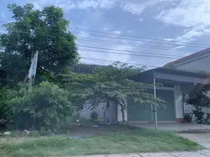 Bức ảnh chụp một khung cảnh ven đường ở một khu vực dân cư. Nổi bật trong ảnh là một ngôi nhà có tường trắng, mái che bằng tôn và cửa cuốn xếp lớp màu xanh lá cây. Xung quanh nhà và ven đường có nhiều cây xanh tươi tốt. Một cột điện bê tông đứng ở bên trái, có một tấm biển bị rách và không thể đọc được nội dung chữ viết. Phía trên, hệ thống dây điện chằng chịt vắt ngang qua bầu trời xanh có mây trắng. Tiền cảnh là một bãi cỏ ven đường. Không có người hay hoạt động cụ thể nào được nhìn thấy trong ảnh.。（日本語訳）。写真は、ある住宅街の道端の風景を撮影したものです。写真の中で目立っているのは、白い壁、トタンの屋根、そして緑色の巻き上げ式シャッタードアを持つ一軒の家です。家の周りや道端には、青々とした緑の木々がたくさんあります。左側にはコンクリートの電柱が立っており、破れて文字の内容が読めない看板が一つあります。上方では、複雑に絡み合った電線が、白い雲のある青空を横切っています。前景は道端の草地です。写真には人や特定の活動は見られません。