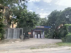 Bức ảnh chụp một cửa hàng xăng dầu ven đường. Trên mái che màu xanh dương của trạm xăng có dòng chữ lớn màu vàng 'H PHÁT PETRO' và phía dưới là biển hiệu 'CỬA HÀNG XĂNG DẦU MINH KHANG'. Có nhiều trụ bơm xăng được đặt trên sân bê tông. Xung quanh trạm xăng được bao bọc bởi nhiều cây xanh tươi tốt. Bên trái là một ngôi nhà nhiều tầng màu vàng với cổng sắt xếp. Bên phải, có một vài chiếc xe máy đang đỗ. Gần lối vào có một chiếc võng được mắc. Trên sân, có một con chim nhỏ màu trắng. Bầu trời có mây. Không có hoạt động mua bán nào đang diễn ra tại thời điểm chụp ảnh.。（日本語訳）。道路沿いのガソリンスタンドを撮影した写真です。ガソリンスタンドの青い屋根には「H PHÁT PETRO」という大きな黄色い文字があり、その下には「CỬA HÀNG XĂNG DẦU MINH KHANG」という看板があります。コンクリートの敷地に多くの給油ポンプが設置されています。ガソリンスタンドの周りは、青々と茂る多くの木々に囲まれています。左側には、折りたたみ式の鉄の門がある黄色い多層階の家があります。右側には、数台のバイクが停まっています。入り口の近くにハンモックが一つ掛けられています。敷地には、一羽の小さな白い鳥がいます。空は曇っています。撮影時には、売買の活動は行われていません。