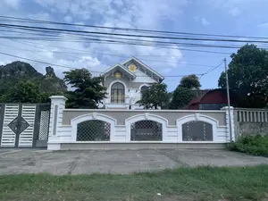 Bức ảnh chụp một ngôi biệt thự lớn, có kiến trúc hiện đại, được sơn màu trắng và xám. Ngôi nhà có nhiều tầng, mái ngói, và ban công với lan can màu vàng. Trên hai đầu hồi của mái nhà có gắn hai biểu tượng hình tròn màu vàng, bên trong là chữ Hán được cách điệu; có thể nhận ra một trong số đó là chữ 'Phúc' (福).。Ngôi nhà được bao quanh bởi một bức tường rào kiên cố, sơn màu trắng và xám, có các ô cửa sổ trang trí với lưới sắt. Cổng chính cũng được thiết kế hiện đại, kết hợp giữa màu trắng và xám.。Bối cảnh phía sau là cây cối xanh tốt và những dãy núi đá vôi. Bầu trời xanh và có nhiều mây. Phía trên của bức ảnh có nhiều đường dây điện và cáp viễn thông chằng chịt. Không có người hay hoạt động nào đang diễn ra trong ảnh.。（日本語訳）。この家は多層階で、瓦屋根、そして黄色い手すりのあるバルコニーがあります。屋根の両方の切妻には、黄色い円形のシンボルが2つ取り付けられており、その中にはデザイン化された漢字があります。そのうちの一つは「福」という字であることが分かります。家は、白とグレーで塗装された頑丈な塀に囲まれており、鉄格子で装飾された窓のような開口部があります。正門も同様にモダンなデザインで、白とグレーを組み合わせています。背景には、青々とした木々と石灰岩の山々があります。空は青く、多くの雲があります。写真の上部には、多くの電線や通信ケーブルが張り巡らされています。写真の中には人や活動の様子は見られません。