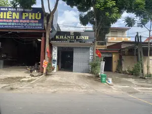 Đây là hình ảnh chụp một con phố tại Việt Nam, có các cửa hàng và nhà xưởng san sát nhau. Bên trái là một cơ sở kinh doanh có biển hiệu lớn màu xanh ghi "NHÀ MÁY TÔN THÉP HIỀN PHÚ", chuyên kinh doanh "TÔN MẠ MÀU - MẠ KẼM - TÔN LẠNH VIỆT - NHẬT SSSC" và "THÉP HỘP U-I-V... PHỤ KIỆN CÁC LOẠI". Địa chỉ được ghi trên biển là "XÃ VĨNH LỘC - HUYỆN VĨNH LỘC, TỈNH THANH HÓA". Bên trong nhà xưởng, có một người đang ngồi làm việc và một chiếc máy cắt sắt. Ở giữa là cửa hàng "KHÁNH LINH STORE" với biển hiệu màu đen, chữ vàng, địa chỉ "SN 208 ĐƯỜNG TRỊNH KHẢ - THỊ TRẤN VĨNH LỘC". Cửa hàng này có cửa xếp kim loại đang mở, bên trong có chữ "Kính Chào". Các biển hiệu dọc cho thấy cửa hàng bán "Thời Trang Nam", "Phụ Kiện", "ĐỒNG HỒ", "GIÀY DÉP". Hai lá cờ Tổ quốc Việt Nam được treo ở phía trước. Dọc vỉa hè có cây xanh, chậu cây cảnh, một thùng rác màu xanh và nhiều dây điện chằng chịt phía trên.。（日本語訳）。これはベトナムのある通りを撮影した写真で、店舗や工場が隣接しています。左側には「NHÀ MÁY TÔN THÉP HIỀN PHÚ」と書かれた大きな青い看板を掲げた事業所があり、「TÔN MẠ MÀU - MẠ KẼM - TÔN LẠNH VIỆT - NHẬT SSSC」（カラーメッキ鋼板 - 亜鉛メッキ鋼板 - ベトナム・日本SSSC製冷延鋼板）と「THÉP HỘP U-I-V... PHỤ KIỆN CÁC LOẠI」（角形鋼管 U-I-V... 各種付属品）を専門に扱っています。看板に記載されている住所は「XÃ VĨNH LỘC - HUYỆN VĨNH LỘC, TỈNH THANH HÓA」（タインホア省ヴィンロック県ヴィンロック社）です。工場の中では、一人が座って作業をしており、鉄の切断機が一台あります。中央には「KHÁNH LINH STORE」という店があり、黒い看板に金色の文字で、住所は「SN 208 ĐƯỜNG TRỊNH KHẢ - THỊ TRẤN VĨNH LỘC」（ヴィンロック町チンカー通り208番地）です。この店は金属製のシャッターが開いており、中には「Kính Chào」（こんにちは/ようこそ）という文字が見えます。縦長の看板には、店が「Thời Trang Nam」（紳士服）、「Phụ Kiện」（アクセサリー）、「ĐỒNG HỒ」（時計）、「GIÀY DÉP」（靴・サンダル）を販売していることが示されています。正面にはベトナム国旗が2枚掲げられています。歩道沿いには緑の木々、鉢植えの観葉植物、緑色のゴミ箱が一つあり、頭上にはたくさんの電線が絡み合っています。