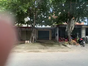 Đây là hình ảnh chụp một con đường với các cửa hàng ven đường. Phía trước có hai cây xanh lớn. Bên trái là một cửa hàng có cửa cuốn đang đóng, trên biển hiệu có ghi 'NẾP NẾP' và 'Tóc - Món Hàng Hịu Kết Đẹp Qu...'. Trên thân cây gần đó có một tấm biển quảng cáo các dịch vụ làm đẹp như 'UỐNG MI' (có thể là Uốn mi), 'TẨY TRANG', 'NỐI MI', 'MASSAGE', cho thấy đây là khu vực có tiệm làm đẹp. Bên phải là một quán nước vỉa hè, nơi một vài người đang ngồi tại bàn ghế nhựa màu đỏ. Cạnh đó có một chiếc xe máy đang đỗ và một đống quả dừa dưới gốc cây. Một phần của bức ảnh ở bên trái bị mờ, có thể do ngón tay che mất.。（日本語訳）。これは、道端に店が並ぶ通りを撮影した写真です。前方には2本の大きな緑の木があります。左側にはシャッターが閉まっている店があり、看板には「NẾP NẾP」と「Tóc - Món Hàng Hịu Kết Đẹp Qu...」と書かれています。近くの木の幹には、「UỐNG MI」（おそらくUốn mi、まつげパーマ）、「TẨY TRANG」（メイク落とし）、「NỐI MI」（まつげエクステ）、「MASSAGE」（マッサージ）といった美容サービスの広告看板があり、この地域に美容院があることを示しています。右側には路上カフェがあり、何人かの人が赤いプラスチック製のテーブルと椅子に座っています。その隣にはバイクが1台停まっており、木の下にはココナッツの山があります。写真の左側の一部はぼやけており、指で隠れてしまったのかもしれません。