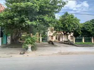 画像は、ある住宅地、おそらくは農村部か郊外の、とある道沿いの光景を記録したものです。写真の中央には、クリーム色に塗られ、緑色の鉄柵で囲まれた2階建ての建物があります。建物の前には、庭へと続く小さな橋があります。入り口の上には、黄色い文字の入った赤い横断幕がありますが、内容は読み取れません。左側には、青いシャッター扉の店と看板がありますが、こちらも内容は不明です。風景には、葉が鬱蒼と茂った大きな緑の木々が多くあります。道端には、鉢植えの観葉植物と黒いプラスチック製のカゴが一つあります。空には白い雲と多くの電線があります。写真には、人の活動は記録されていません