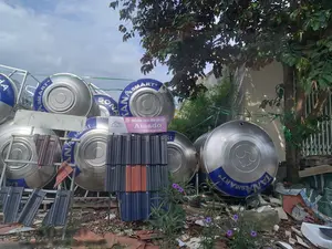 Bức ảnh chụp một khu vực ngoài trời, có vẻ là nơi lưu trữ hoặc trưng bày vật liệu xây dựng. Nổi bật trong ảnh là nhiều bồn nước inox lớn, hình trụ, được xếp chồng lên nhau trên các kệ sắt. Trên các bồn nước có in chữ 'SƠN HÀ' và 'SMART+'. Ở trung tâm, có các chồng ngói lợp với nhiều màu sắc khác nhau như đen, nâu đỏ và xanh dương. Phía trước chồng ngói là một tấm biển quảng cáo màu hồng và trắng có ghi: logo 'D Amado', 'KỶ NGUYÊN NGÓI GỐM TRÁNG MEN CAO CẤP', 'Amado', và khẩu hiệu 'Xây giá trị dựng ước mơ'. Nền đất lộn xộn với các mảnh vỡ của gạch ngói cũ. Bên phải có một cây xanh lớn, và một vài bông hoa dại màu tím mọc lên. Phía sau là một bức tường và bầu trời có mây.。（日本語訳）。写真は屋外のあるエリアを撮影したもので、建築資材の保管場所または展示場のようです。写真の中で目立っているのは、鉄製の棚の上に積み重ねられた、多くの大きな円筒形のステンレス製水タンクです。水タンクには「SƠN HÀ」と「SMART+」という文字が印刷されています。中央には、黒、赤褐色、青など、さまざまな色の屋根瓦の山があります。瓦の山の前にはピンクと白の広告看板があり、そこには「D Amado」のロゴ、「高級釉薬陶器瓦の時代」、「Amado」、そして「価値を築き、夢を建てる」というスローガンが書かれています。地面は古いレンガや瓦の破片で散らかっています。右側には大きな緑の木があり、紫色の野花が数輪咲いています。後方には壁と曇り空が見えます。