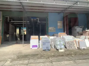 Đây là hình ảnh mặt tiền của một cửa hàng vật liệu xây dựng, chuyên bán gạch ốp lát. Phía trước cửa hàng, trên nền bê tông, có nhiều chồng gạch với các mẫu mã, màu sắc và kích thước đa dạng được trưng bày để bán. Một số mẫu gạch được đặt trên các giá đỡ nhỏ. Bên trong cửa hàng, có thể thấy thêm các kệ trưng bày gạch và các tấm vật liệu khác. Cửa hàng có mái che bằng tôn. Trên tường có treo các bảng mẫu gạch nhưng không thể đọc rõ nội dung. Không có người xuất hiện trong ảnh, chỉ có các sản phẩm được trưng bày.。（日本語訳）。これは、タイルを専門に販売する建材店の正面の画像です。店の前、コンクリートの地面の上には、様々なデザイン、色、サイズのたくさんのタイルの山が販売用に陳列されています。いくつかのタイルサンプルは、小さなスタンドの上に置かれています。店内には、さらにタイルの陳列棚やその他の板材が見えます。店にはトタン製の屋根があります。壁にはタイルのサンプルボードが掛けられていますが、内容ははっきりと読み取れません。写真には誰も写っておらず、陳列された商品だけがあります。