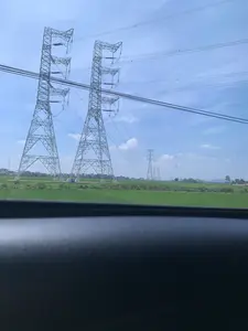 Bức ảnh chụp một khung cảnh nông thôn, nhìn từ bên trong một phương tiện di chuyển, có thể là ô tô. Cảnh vật chính là một cánh đồng lúa xanh rộng lớn, trên đó có một hệ thống đường dây điện cao thế. Nhiều cột điện cao thế bằng kết cấu thép lớn đứng sừng sững giữa cánh đồng, nối tiếp nhau chạy dài về phía chân trời. Các đường dây điện vắt ngang qua bầu trời xanh và có mây trắng. Ở phía xa, có thể thấy lác đác nhà cửa và cây cối. Dưới chân một cột điện ở phía trước, có một vài người và một tấm bạt màu xanh, có thể họ là công nhân đang làm việc hoặc nông dân đang nghỉ ngơi. Không có chữ viết hay biển báo nào có thể đọc được trong ảnh.。（日本語訳）。写真は、ある田園風景を写しており、車内のような移動手段の中から撮影されたものです。主な景色は広大な緑の田んぼで、その上には高圧送電線のシステムがあります。多くの大きな鉄骨構造の高圧鉄塔が田んぼの真ん中にそびえ立ち、地平線に向かって長く連なっています。送電線は青空と白い雲を横切っています。遠くには、家や木々がまばらに見えます。手前にある鉄塔の麓には、数人の人々と青いシートがあり、彼らは作業中の作業員か、休憩中の農家の人々かもしれません。写真の中には、読み取れる文字や標識はありません。