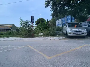 Bức ảnh chụp một khung cảnh ven đường. Trên mặt đường nhựa có vạch kẻ ô màu vàng, có thể là khu vực dành cho xe buýt dừng đỗ. Một chiếc ô tô 7 chỗ màu bạc đang đỗ ở lề đường. Phía sau chiếc xe là một khu đất có nhiều cây cối và bụi rậm. Có một số biển hiệu và biển quảng cáo có thể đọc được nội dung như sau:。1. Biển báo điểm dừng xe buýt: "TUYẾN SỐ 02", "ĐIỂM DỪNG XE BUÝT".。2. Biển hiệu màu trắng của công ty xi măng: "XUAN THANH GROUP", "XI MĂNG XUÂN THÀNH".。3. Biển quảng cáo màu xanh dương: "CÔNG TY TNHH TM SỐ 1.", "TỔNG ĐÀI TAXI THANH HÓA".。4. Chữ trên tòa nhà màu vàng ở phía xa: "TỔNG CÔNG TY XI MĂNG VIỆT NAM", "NHÀ PHÂN PHỐI XI MĂNG BỈM SƠN".。Khung cảnh chung cho thấy đây là một khu vực ven đường, có thể gần các khu công nghiệp hoặc cơ sở kinh doanh vật liệu xây dựng tại Thanh Hóa.。（日本語訳）。写真は道端の風景を写したものです。アスファルトの路上には黄色の格子状の線が引かれており、バスの停車エリアかもしれません。銀色の7人乗り自動車が路肩に停まっています。車の後ろには、多くの木々や茂みがある土地が広がっています。いくつかの看板や広告があり、その内容は次のように読み取れます。1. バスの停留所標識：「02番路線」、「バス停留所」。2. セメント会社の白い看板：「XUAN THANH GROUP」、「XUAN THANHセメント」。3. 青い広告看板：「第1商事有限会社」、「タインホアタクシー配車センター」。4. 遠くにある黄色い建物上の文字：「ベトナムセメント総公社」、「BIM SONセメント販売代理店」。全体の風景から、ここはタインホア省の工業団地や建設資材販売店の近くにある道端のエリアであることがわかります。