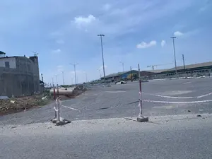 Hình ảnh cho thấy một khu vực rộng lớn đang trong quá trình xây dựng, có thể là một con đường mới hoặc một bãi đậu xe. Bề mặt được rải sỏi đá. Ở bên trái là một ngôi nhà hoặc công trình đang xây dựng dở dang bằng bê tông. Dọc theo khu vực này là hàng loạt cột đèn đường đã được lắp đặt. Ở phía xa, có thể nhìn thấy các tòa nhà lớn giống như nhà xưởng hoặc kho bãi và một chiếc cần cẩu xây dựng. Ở tiền cảnh, có các cọc rào tạm được nối với nhau bằng dây băng cảnh báo màu đỏ và trắng, có thể để ngăn người và phương tiện đi vào khu vực thi công. Bên lề đường còn có một số ống cống bê tông và đất đá xây dựng. Bầu trời trong xanh và có vài gợn mây.。（日本語訳）。写真は、建設中の広大なエリアを写しており、新しい道路か駐車場である可能性があります。表面は砂利が敷き詰められています。左側には、コンクリートでできた、建設途中の家屋か建物があります。このエリアに沿って、多数の街灯が設置済みです。遠くには、工場か倉庫のような大きな建物と、建設用クレーンが見えます。手前には、赤と白の警告テープで結ばれた仮設の杭があり、人や車両が工事区域に立ち入るのを防ぐためのものと思われます。道端には、コンクリート製の土管や建設用の土砂もいくつかあります。空は青く澄んでおり、雲が少し浮かんでいます。